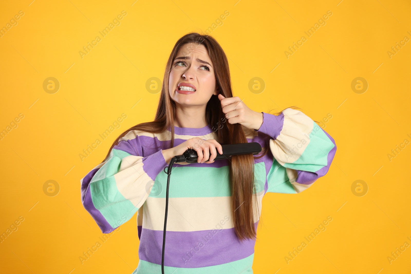 Photo of Stressed young woman with flattening iron on yellow background. Hair damage