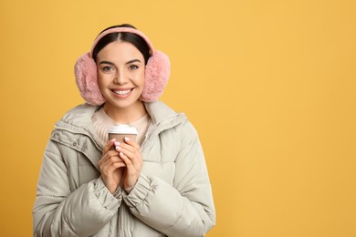 Photo of Beautiful young woman in earmuffs with cup of drink on yellow background. Space for text