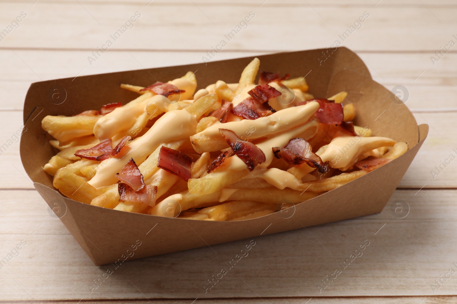 Photo of Tasty potato fries, cheese sauce and bacon in paper container on light wooden table, closeup