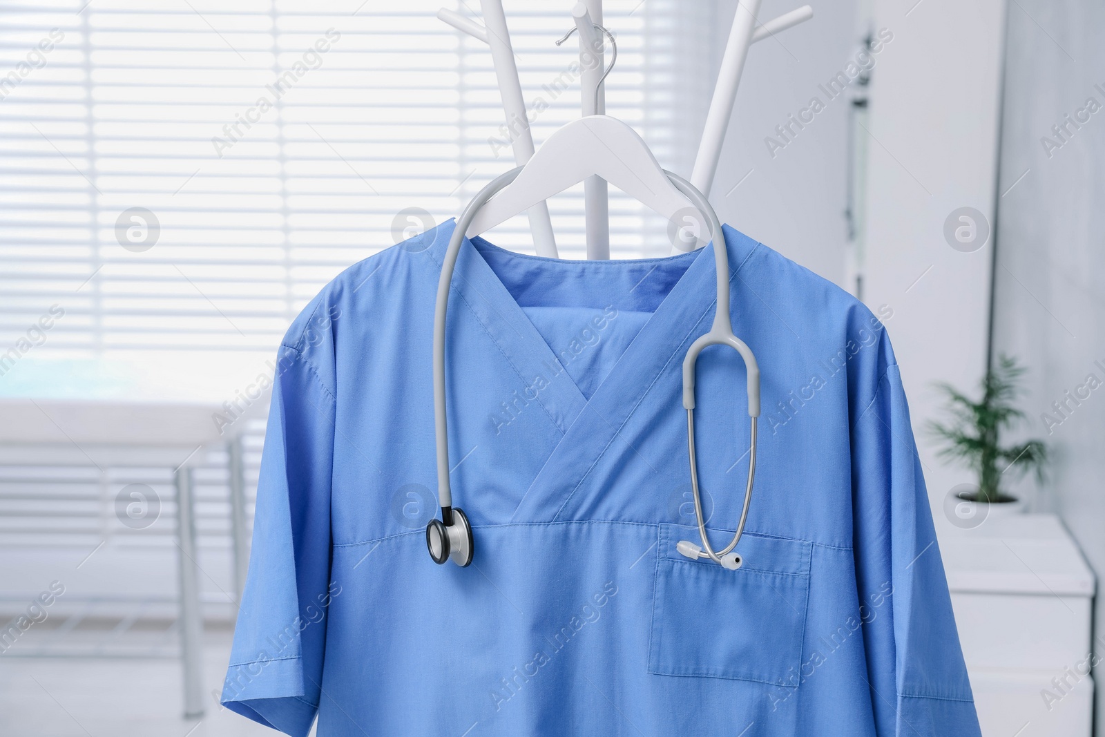 Photo of Blue medical uniform and stethoscope hanging on rack in clinic, closeup