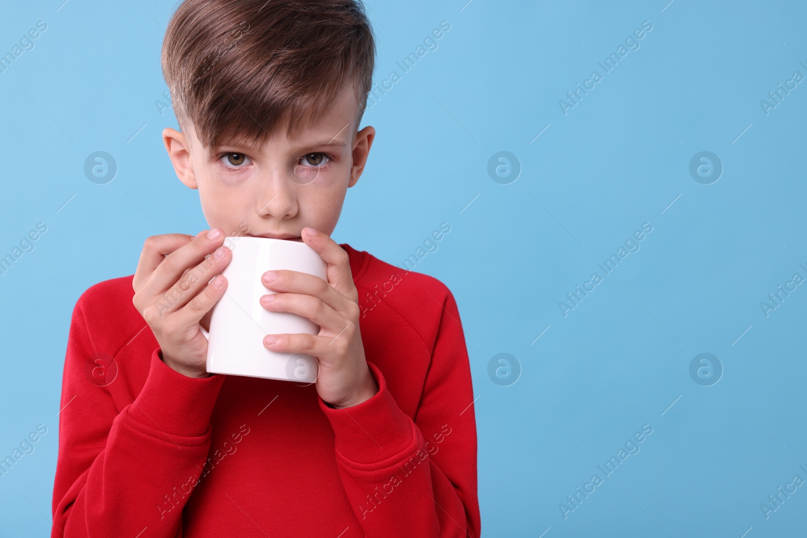 Photo of Cute boy drinking beverage from white ceramic mug on light blue background, space for text