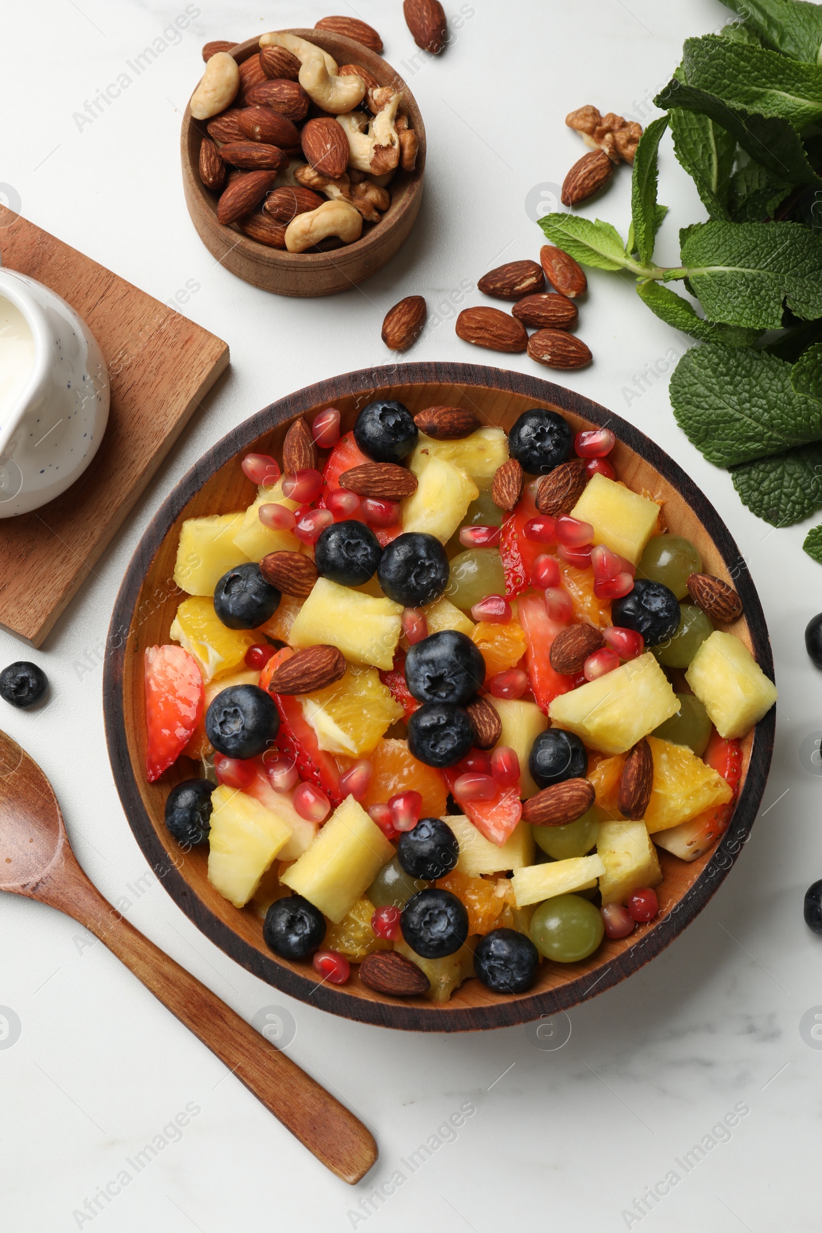 Photo of Delicious fruit salad in bowl, berries, fresh mint and nuts on white marble table, flat lay