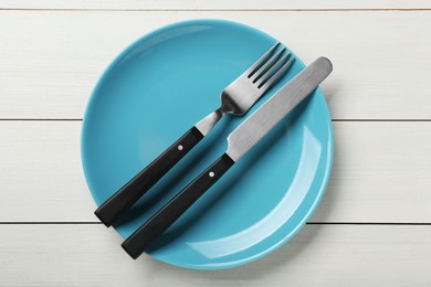 Light blue ceramic plate with cutlery on white wooden table, top view