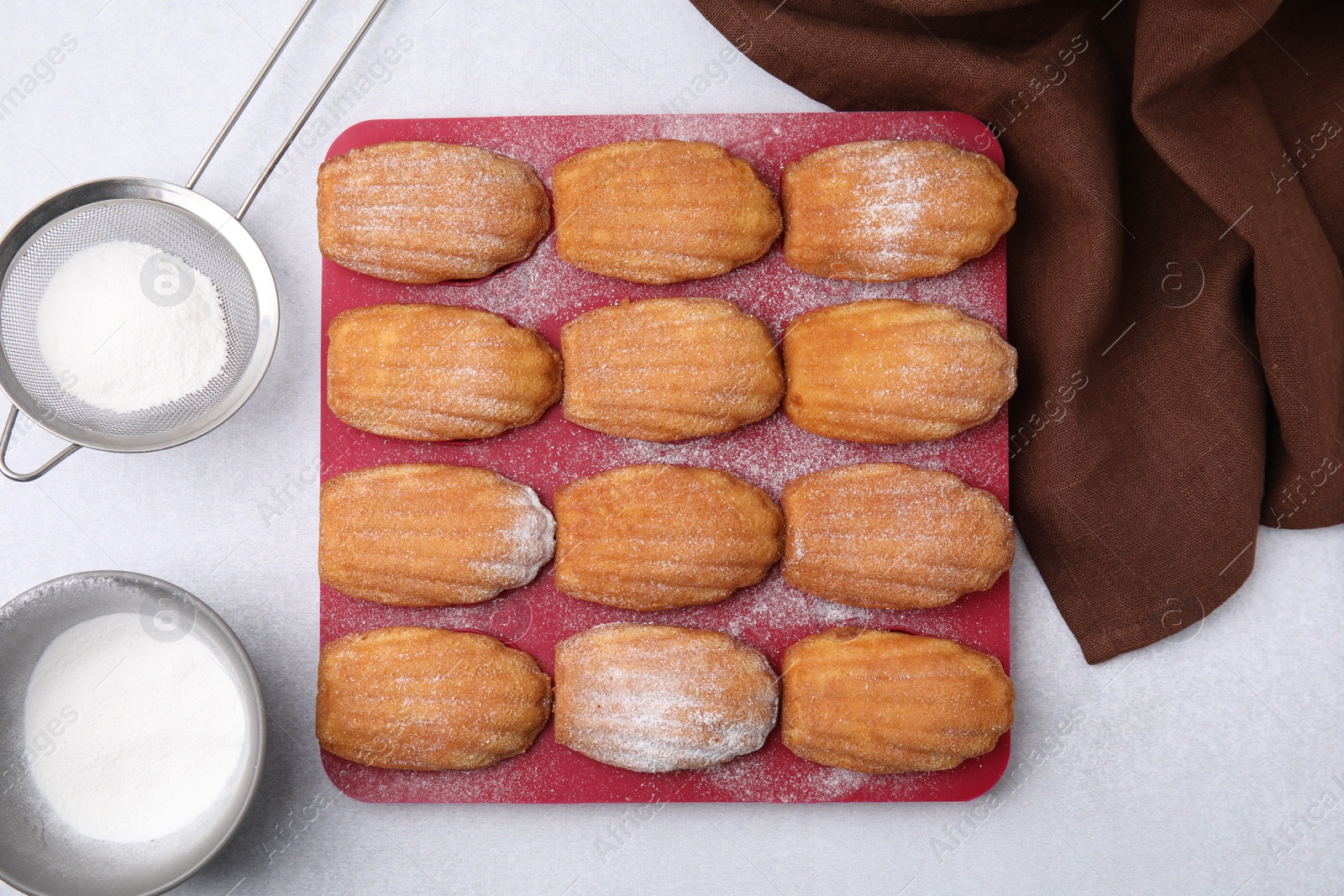 Photo of Delicious madeleine cookies in baking mold and sieve with powdered sugar on white table, flat lay