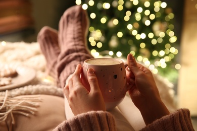 Photo of Woman holding cup of delicious hot drink near Christmas tree indoors, closeup