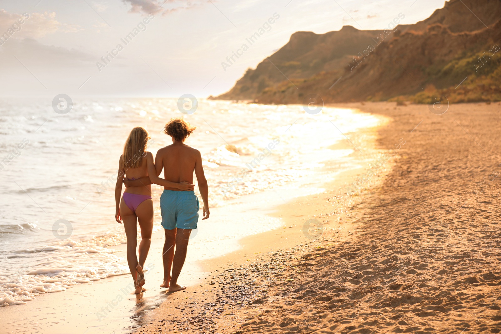 Photo of Young woman in bikini and her boyfriend walking on beach at sunset. Lovely couple