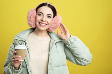 Photo of Beautiful young woman in earmuffs with cup of drink on yellow background