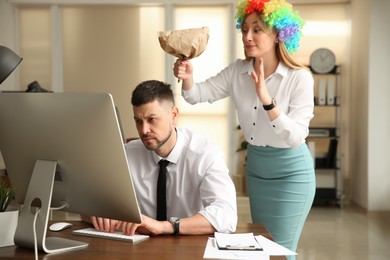 Photo of Young woman popping paper bag behind her colleague in office. Funny joke