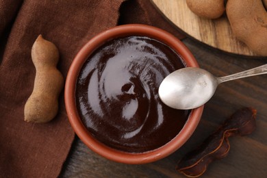 Photo of Bowl of tamarind sauce, spoon and fresh pods on wooden table, flat lay