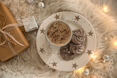 Photo of Tasty hot drink, cookies, gift and Christmas lights on fur, flat lay