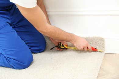 Photo of Worker with cutter knife and measuring tape installing new carpet indoors, closeup