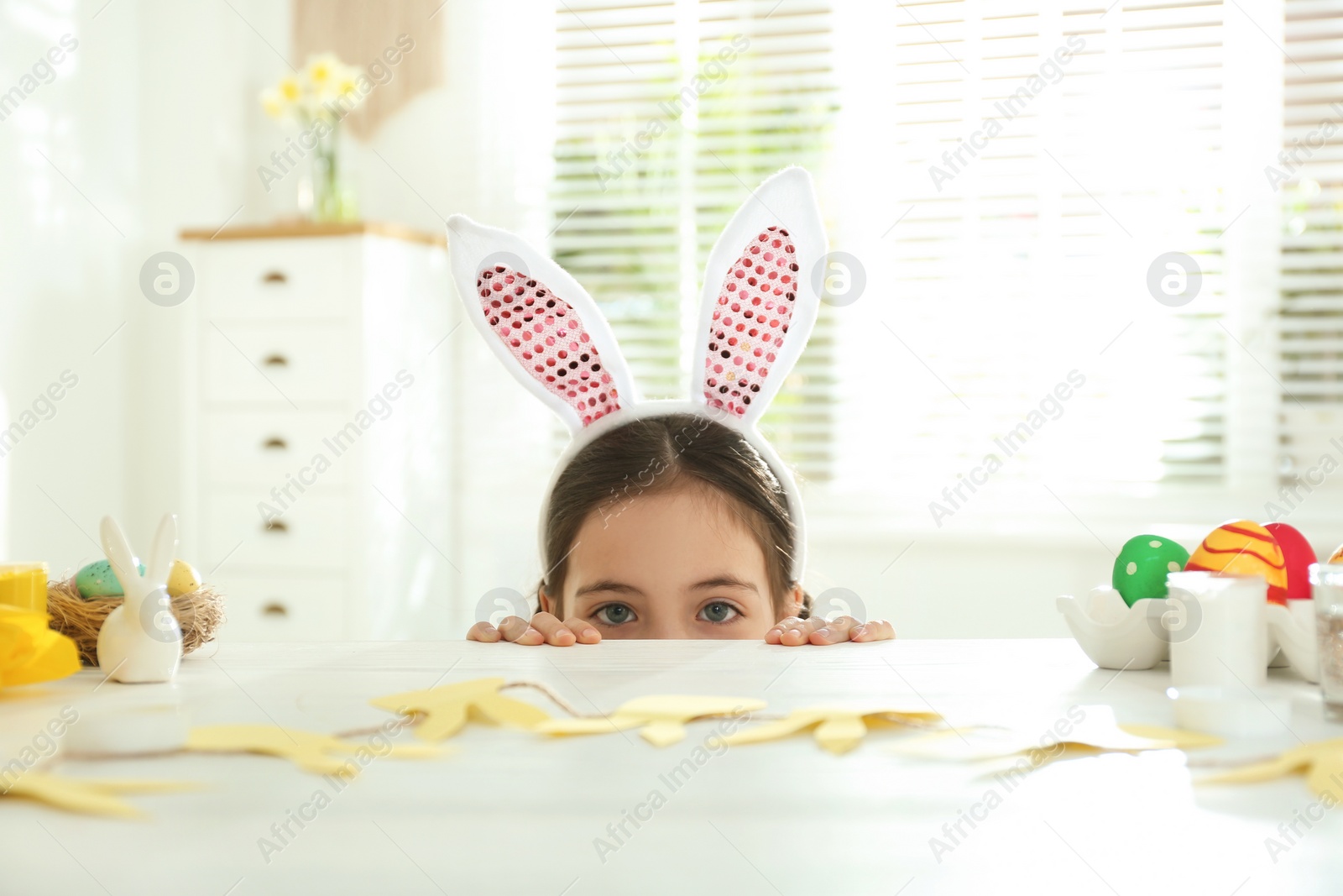 Photo of Cute little girl wearing bunny ears headband at table with Easter eggs, indoors