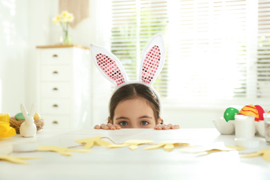 Photo of Cute little girl wearing bunny ears headband at table with Easter eggs, indoors