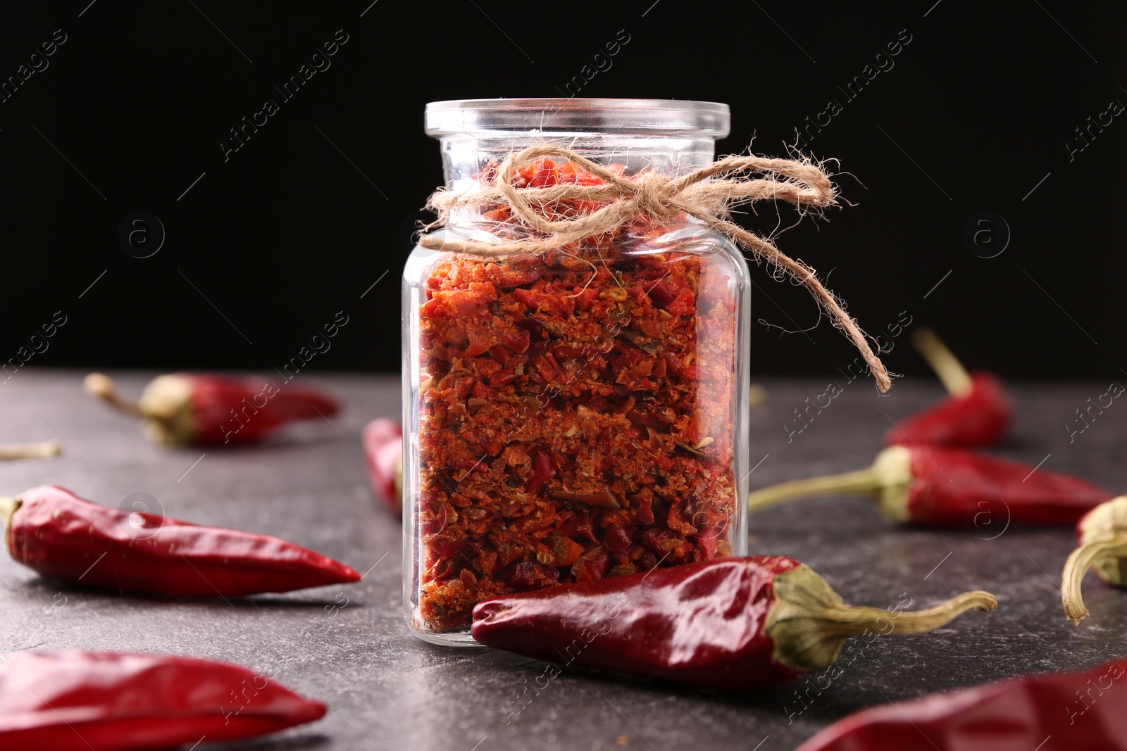 Photo of Chili pepper flakes in jar and pods on textured table against black background, closeup