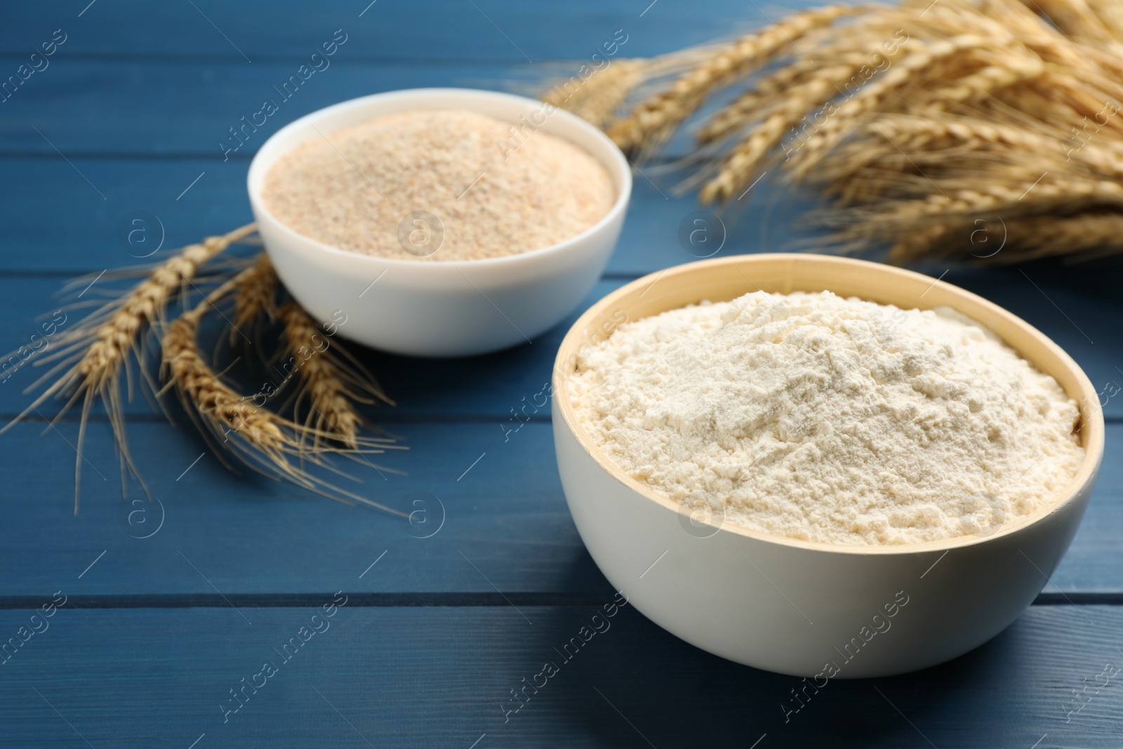 Photo of Bowls of flours and wheat ears on blue wooden table