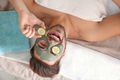 Photo of Young man with clay mask on his face holding cucumber slices in spa salon, above view