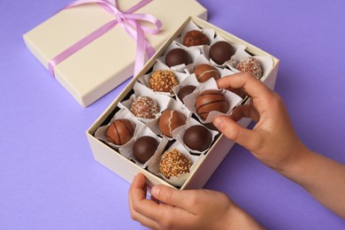 Child with box of delicious chocolate candies on light purple background, closeup