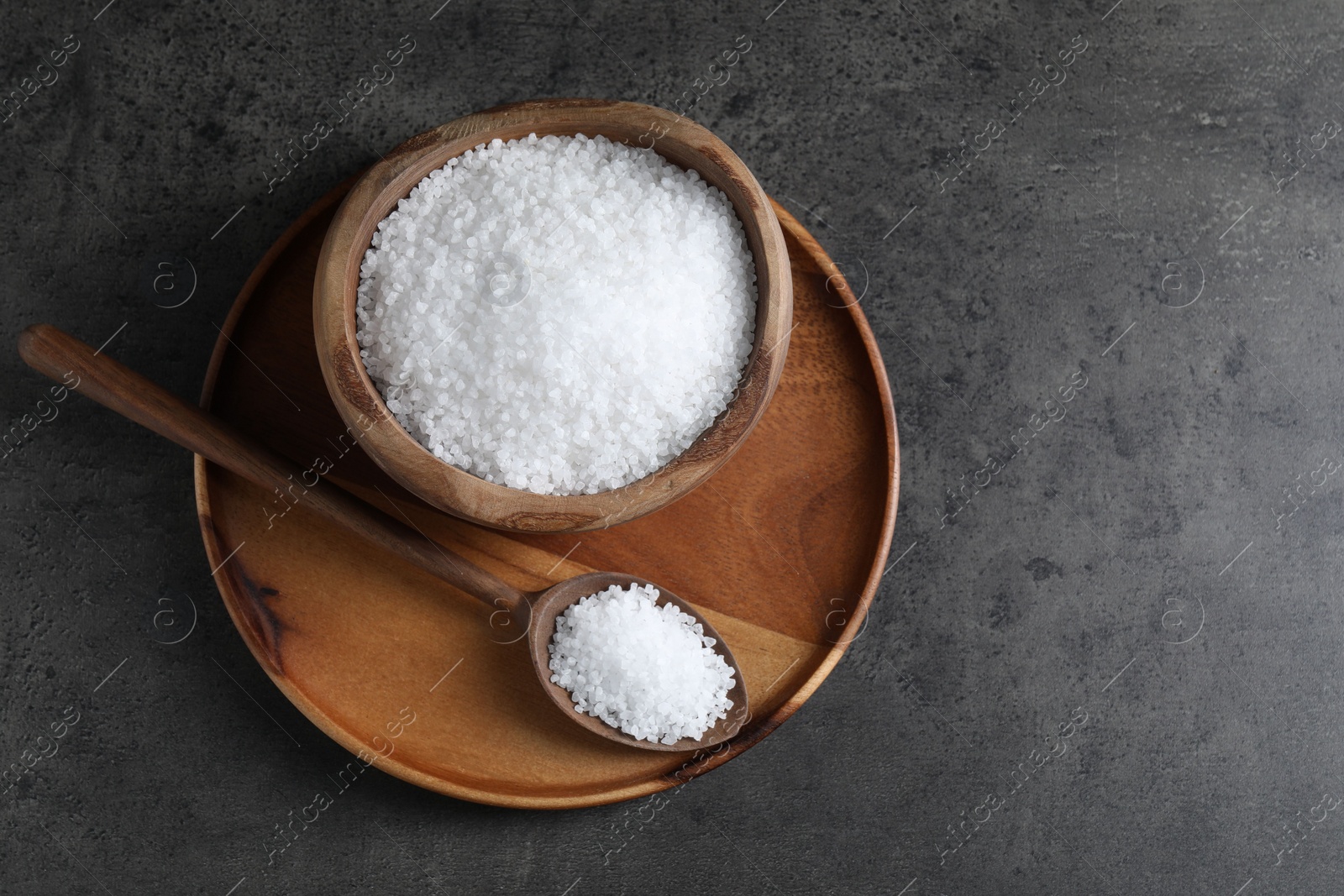 Photo of Natural salt in wooden bowl and spoon on dark grey table, top view. Space for text