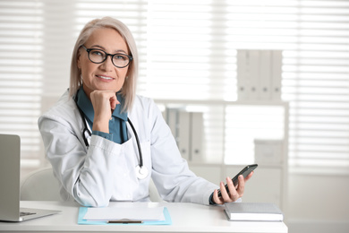 Mature female doctor with smartphone at table in office