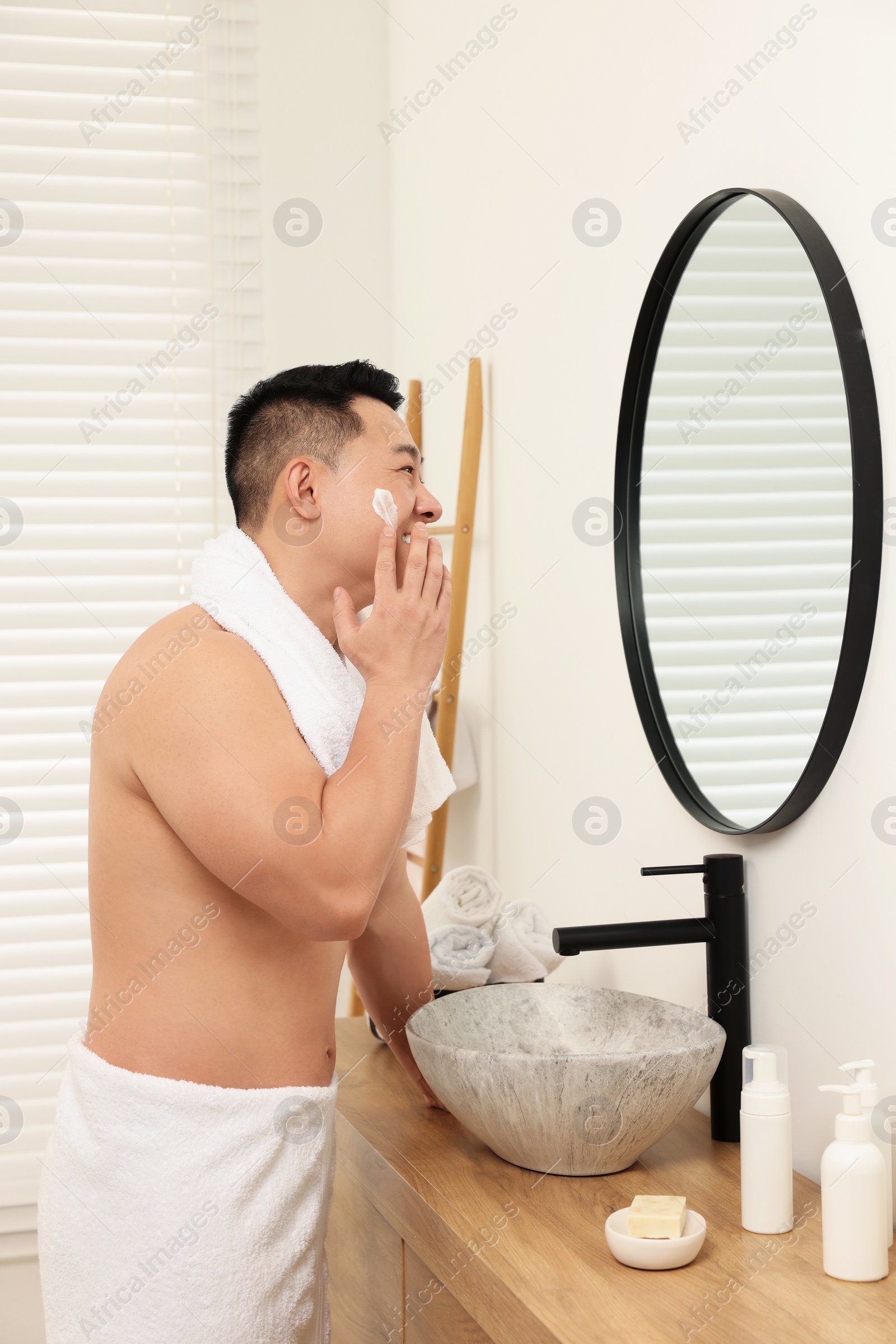 Photo of Handsome man applying cream onto his face near mirror in bathroom