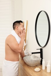 Photo of Handsome man applying cream onto his face near mirror in bathroom