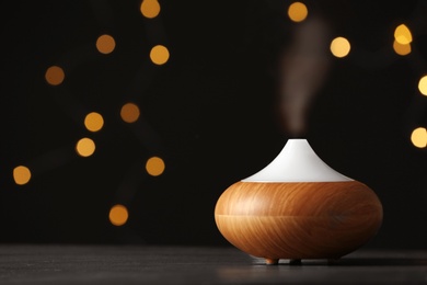 Photo of Modern essential oil diffuser on wooden table against blurred lights, space for text