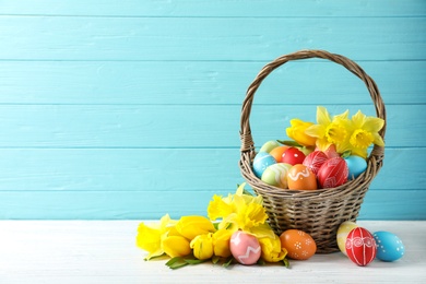 Wicker basket with painted Easter eggs and flowers on table against color background, space for text