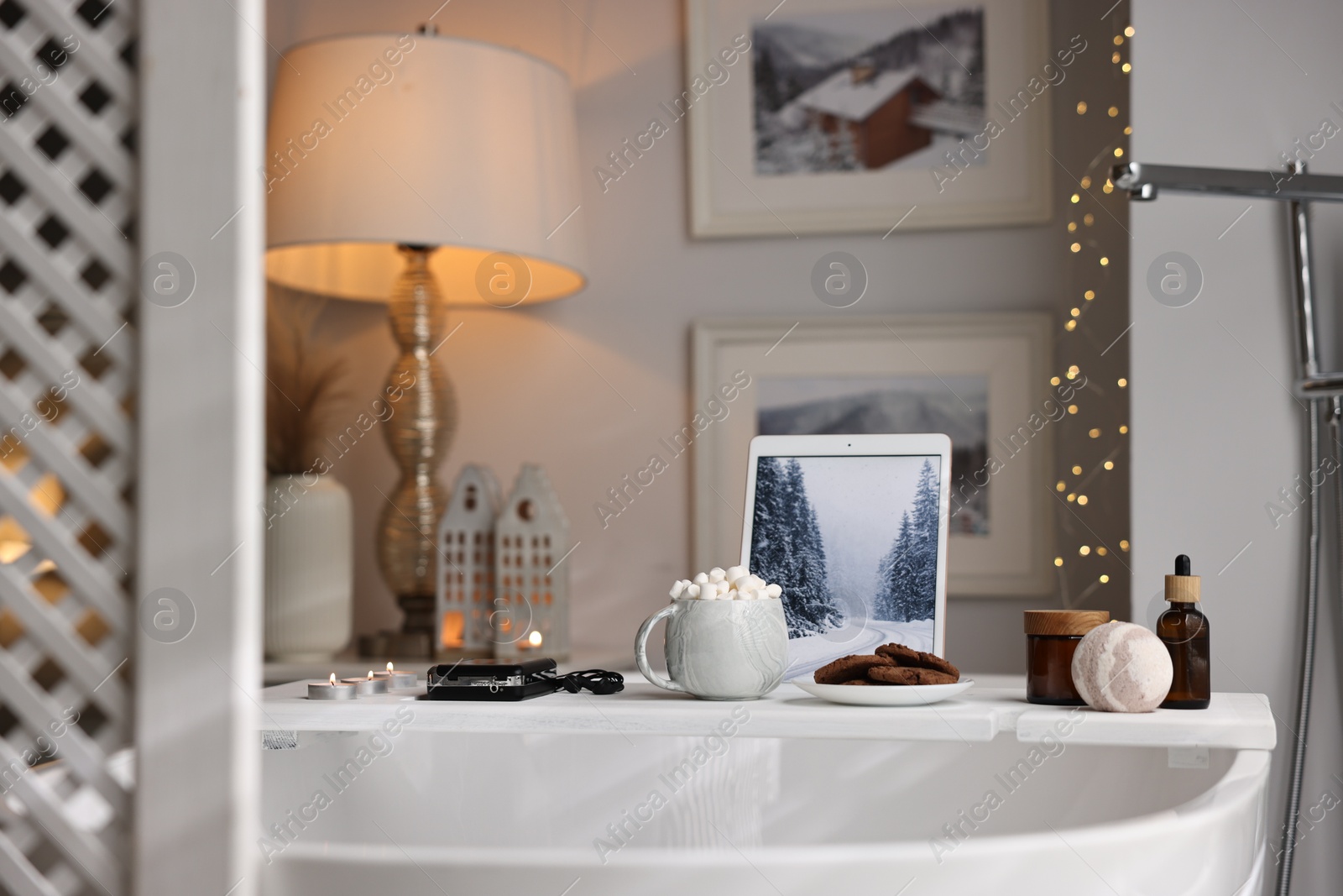 Photo of White wooden tray with tablet, spa products and burning candles on bathtub in bathroom