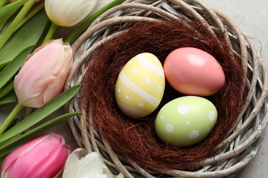Wicker nest with painted Easter eggs and tulips on color background, top view