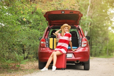 Photo of Happy woman sitting in car trunk with suitcases near forest
