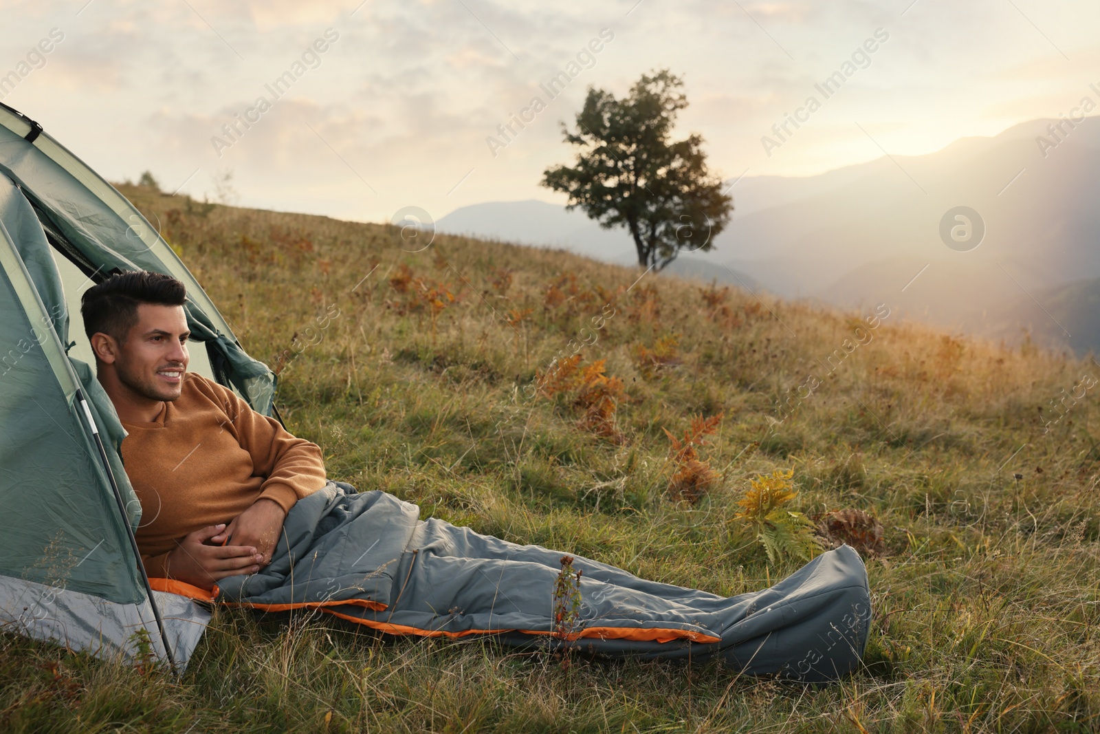 Photo of Man in sleeping bag near camping tent on hill, space for text