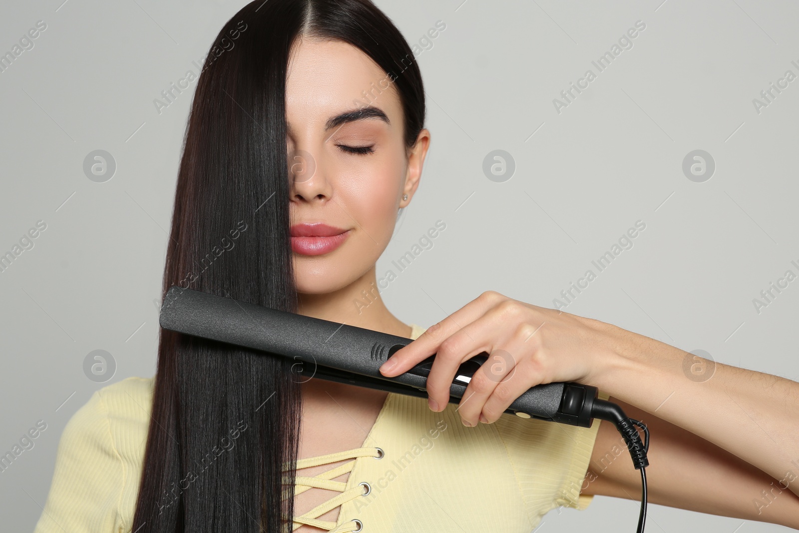 Photo of Beautiful woman using hair iron on light grey background