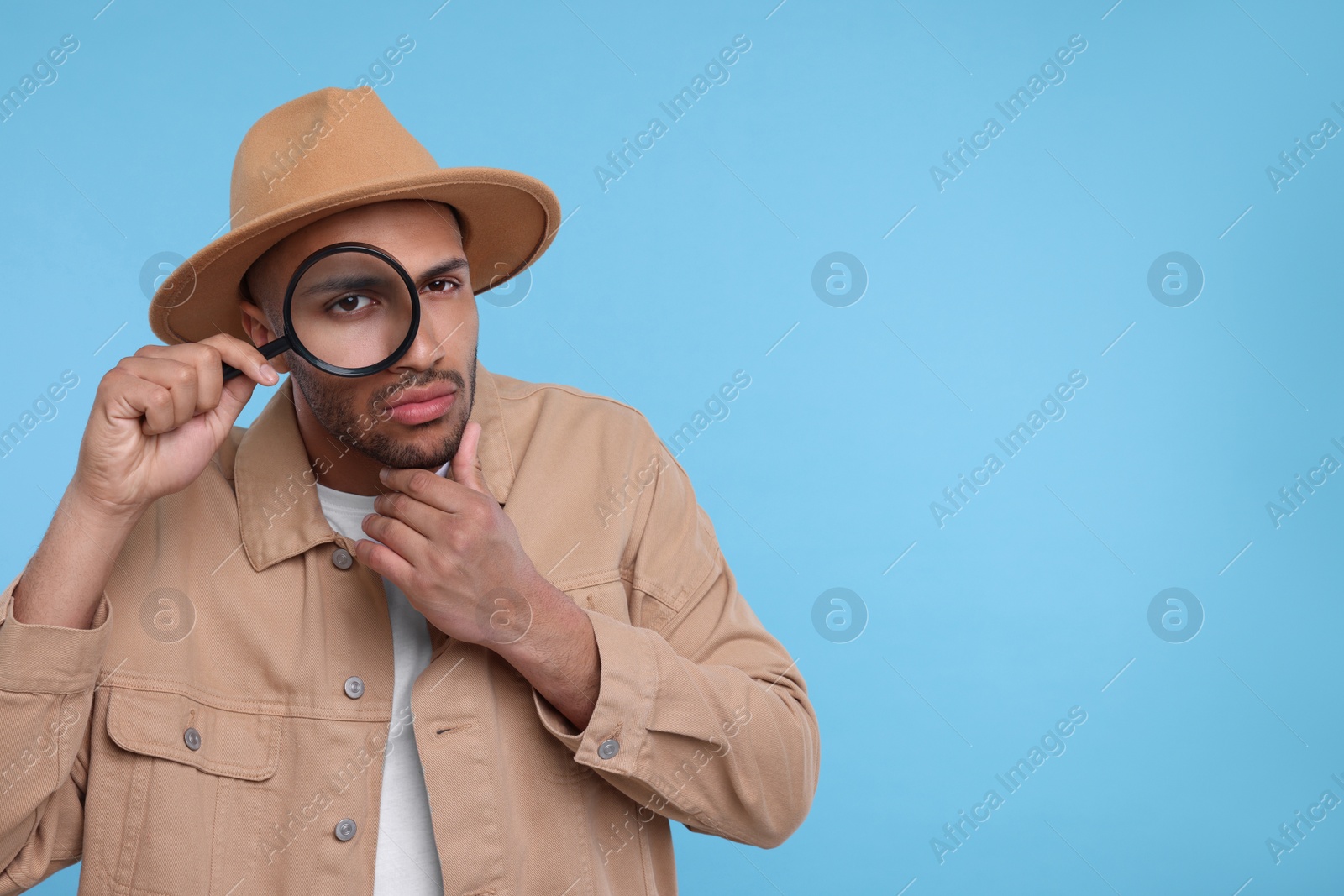 Photo of Curious man looking through magnifier glass on light blue background, space for text