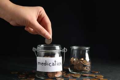 Photo of Woman putting coin into glass jar with tag MEDICATION on black table, closeup