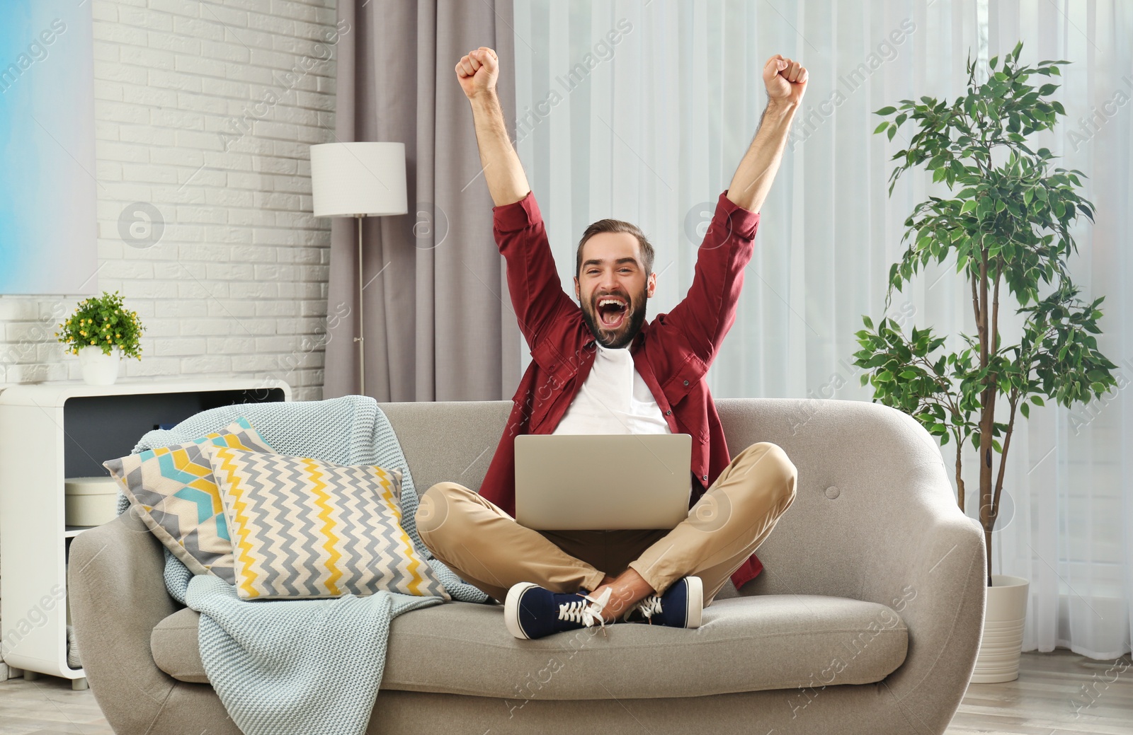 Photo of Emotional young man with laptop celebrating victory on sofa at home