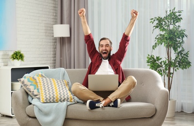 Emotional young man with laptop celebrating victory on sofa at home