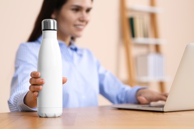 Photo of Young woman taking thermo bottle at workplace indoors, focus on hand