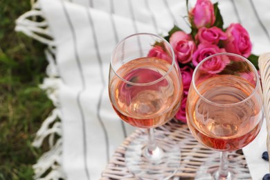 Glasses of delicious rose wine, flowers and basket on picnic blanket outdoors, closeup