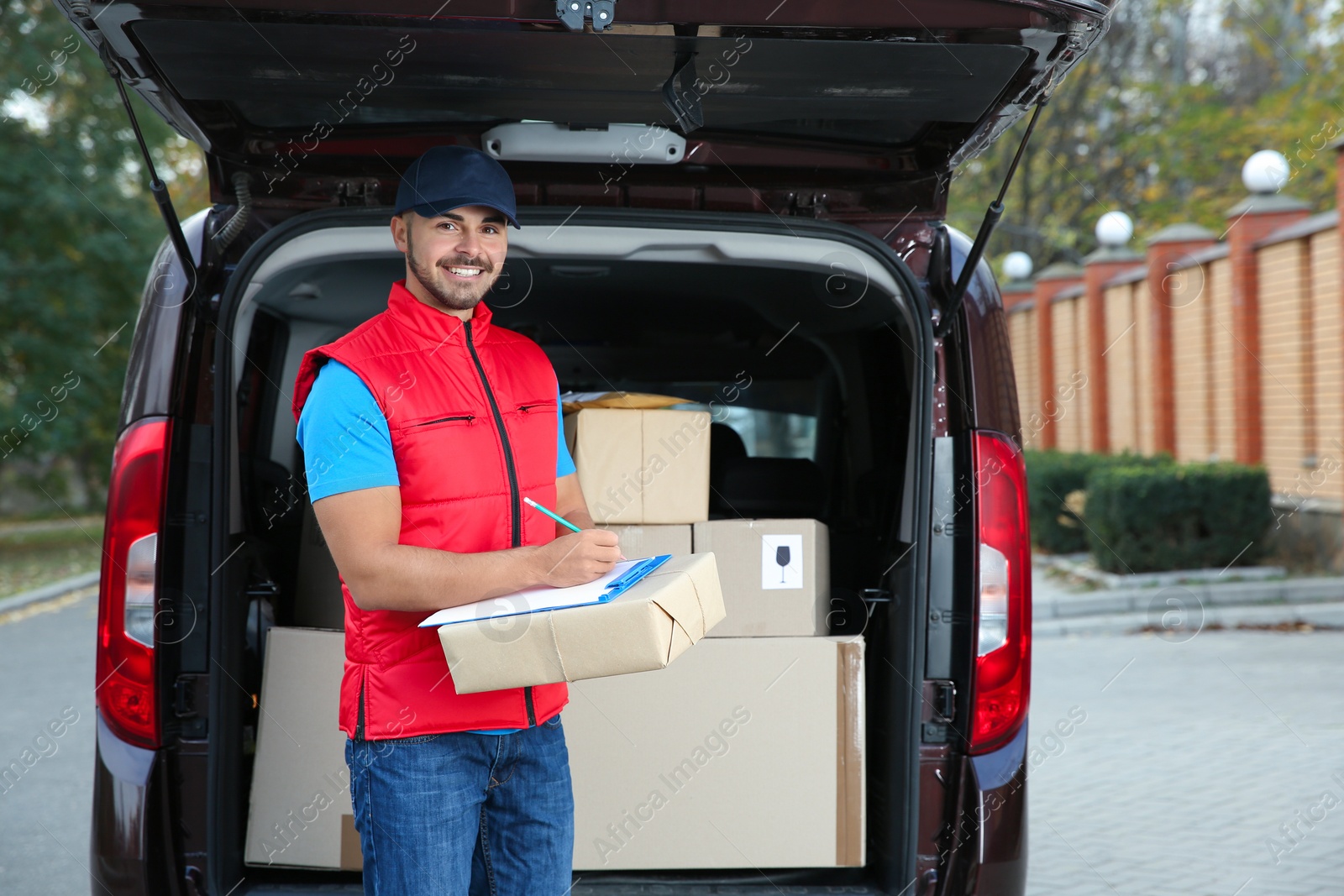 Photo of Young courier with parcel and clipboard near delivery van outdoors