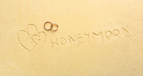 Photo of Word Honeymoon written on sand, hearts two golden rings, top view