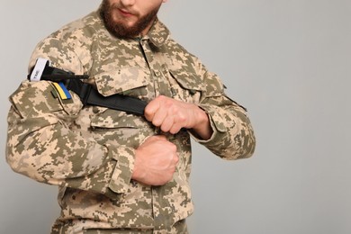 Photo of Ukrainian soldier in military uniform applying medical tourniquet on arm against light grey background, closeup. Space for text