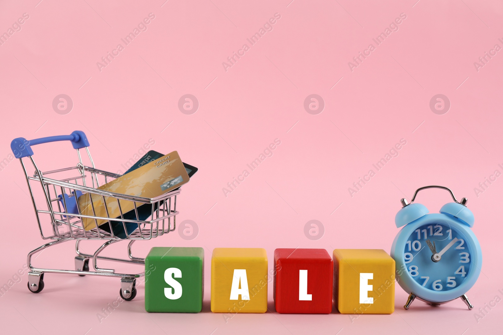 Photo of Word Sale made with colorful cubes, alarm clock and credit cards in shopping cart on pink background
