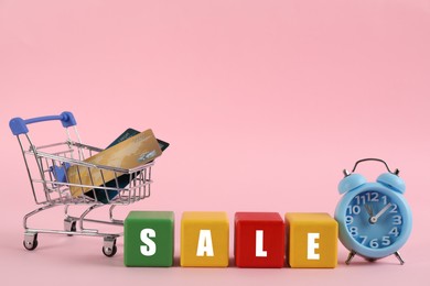 Word Sale made with colorful cubes, alarm clock and credit cards in shopping cart on pink background