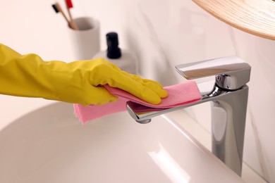 Woman in gloves cleaning faucet of bathroom sink with rag, closeup