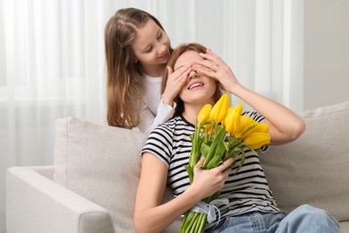 Daughter covering mother's eyes with her palms and congratulating with bouquet of yellow tulips at home