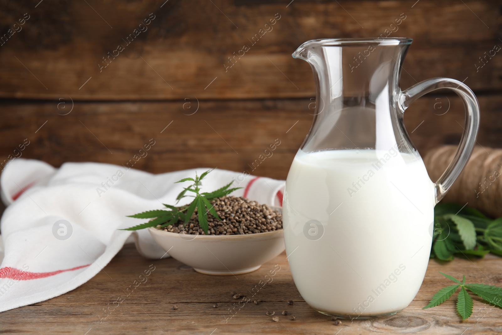 Photo of Glass jug of fresh hemp milk, seeds and leaves on wooden table. Space for text
