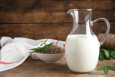 Photo of Glass jug of fresh hemp milk, seeds and leaves on wooden table. Space for text