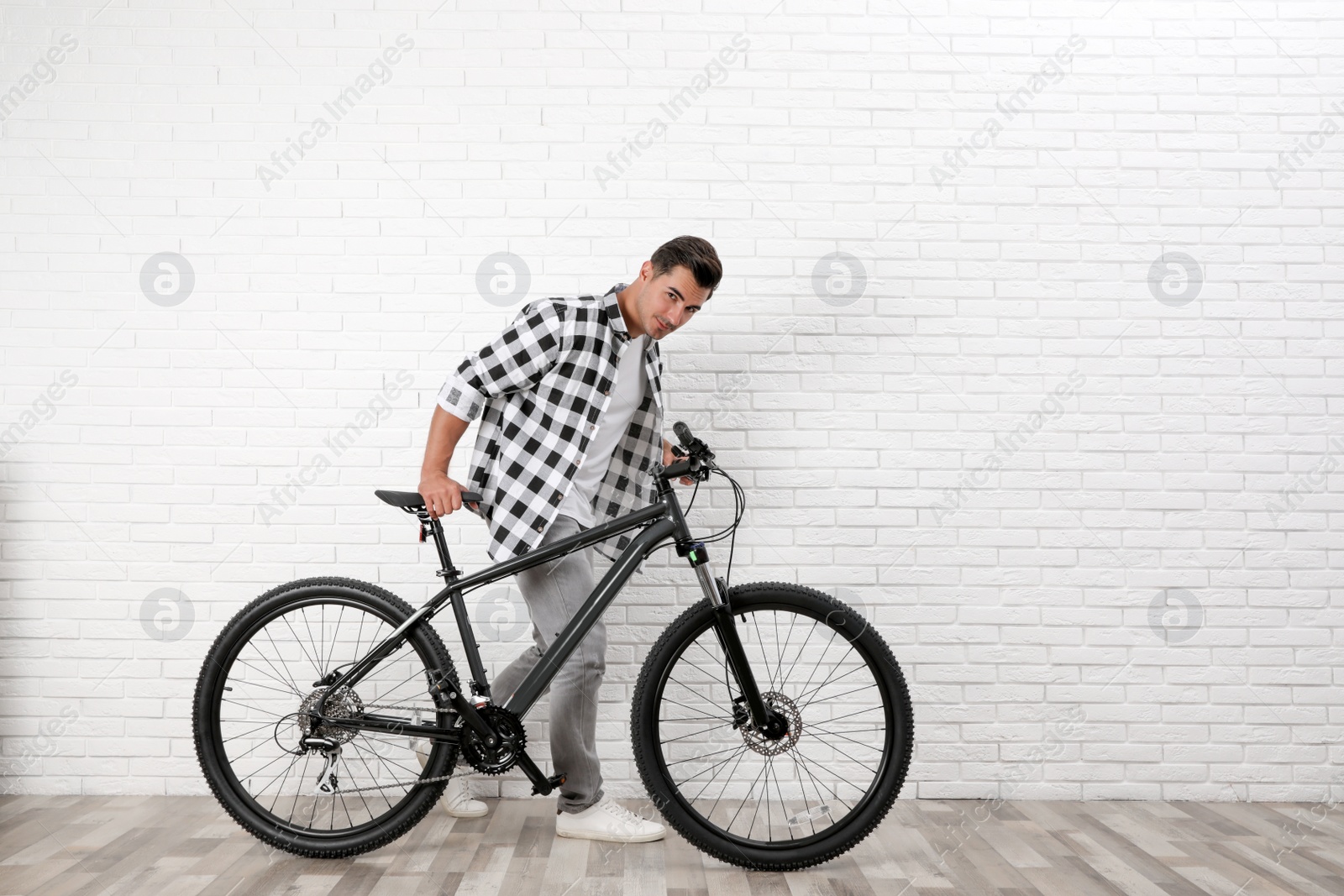 Photo of Handsome young man with modern bicycle near white brick wall indoors