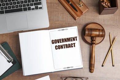 Image of Notebook with words Government Contract on wooden table. Workplace with laptop, stationery and gavel, flat lay