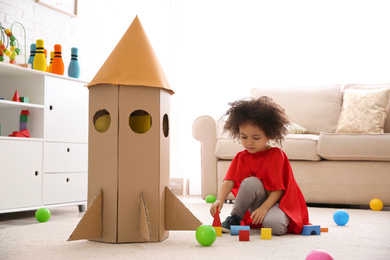Cute African American child playing with cardboard rocket at home